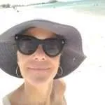 Mulher de cabelos escuros em pé perto do mar, com um sorriso caloroso no rosto.
